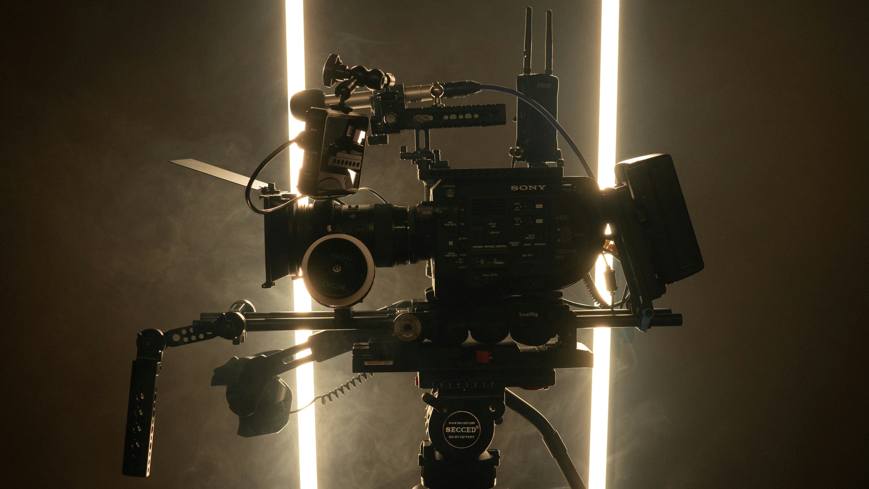 A rigged cinema camera on a tripod, backlit by two tube lights in warm haze