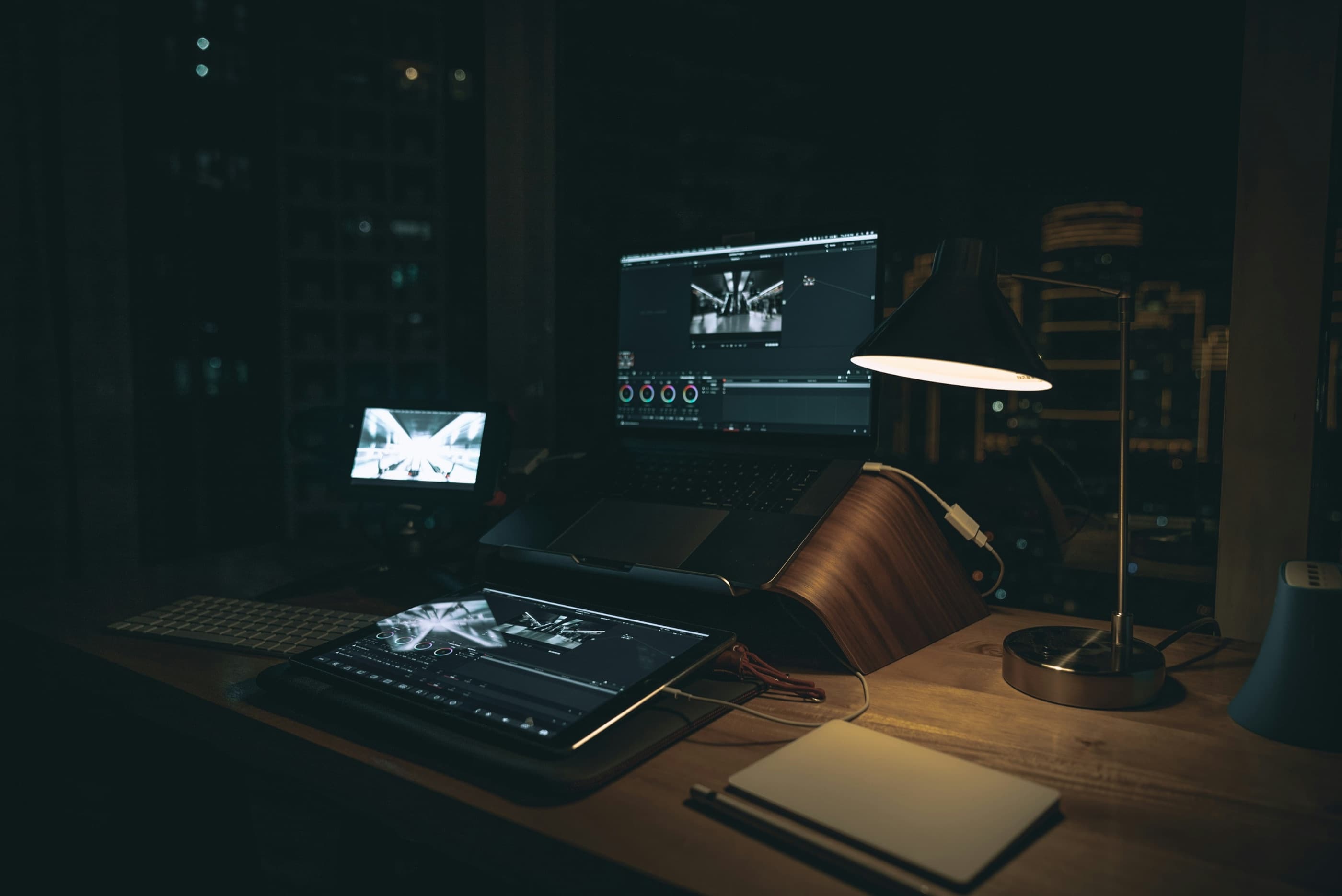 An editing desk at night: a laptop and a tablet running a colour grading app, lit by a warm desk lamp